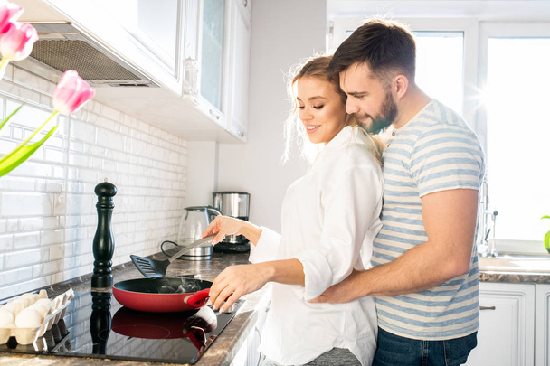 Vue de côté portrait d'un jeune couple aimant cuisiner le petit déjeuner ensemble embrassant doucement dans la cuisine éclairée par la lumière du soleil, espace de copie
 - Photo, image