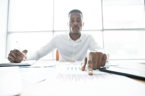 ernsthafter junger afrikanisch-amerikanischer Finanzberater zeigt auf Grafik, während er Papiere in modernem Büro prüft - Foto, Bild