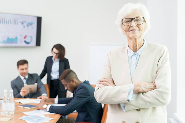 Content confident attractive senior chief executive officer in glasses wearing jacket standing in meeting room and looking at camera while business analysts working with data in background. - Photo, Image