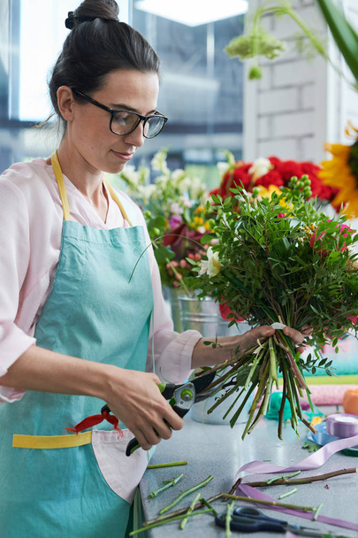 Vista lateral retrato de florista feminina vestindo avental arranjar buquês na loja de flores e sorrindo
 - Foto, Imagem