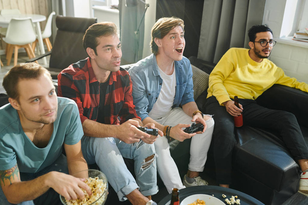 Два хлопці тримають джойстики і борються за перемогу в відеоіграх, а їхні друзі їдять попкорн і п'ють пиво на дивані
 - Фото, зображення