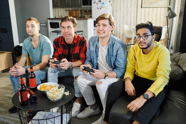Молодий чоловік програє своєму другові в відеогрі, а інші друзі сидять на дивані і дивляться гру
 - Фото, зображення