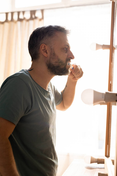 Mañana del hombre adulto: hombre barbudo serio en camiseta de pie delante del espejo con bombillas y cepillarse los dientes en el baño
 - Foto, imagen