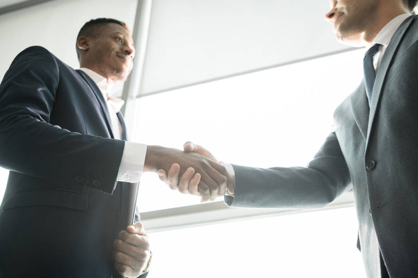 Below view of content modern multi-ethnic business partners in formal suits standing by window and shaking hands after concluding deal - Photo, Image