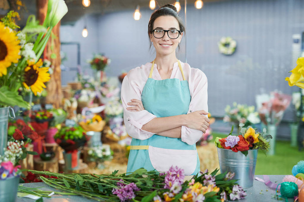 Талия портрет улыбающейся продавщицы позирует с скрещенными руками и смотрит в камеру в маленьком цветочном магазине, копирует пространство
 - Фото, изображение