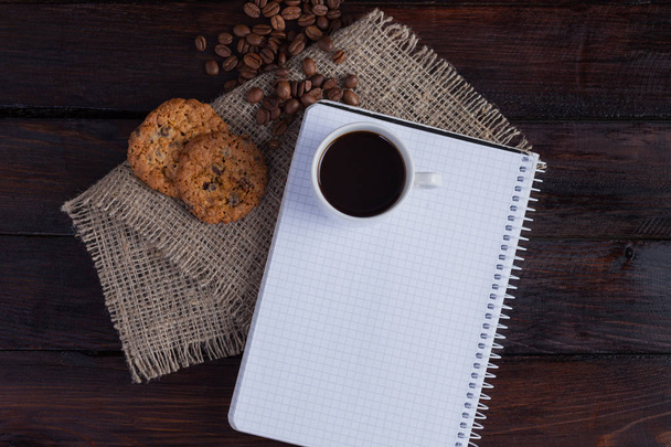 Bílý šálek kávy s drobečky kávy poblíž a sušenky a Poznámkový blok v kleci pro záznamy na lněné tkaniny na tmavém pozadí vinobraní. Pohled shora - Fotografie, Obrázek