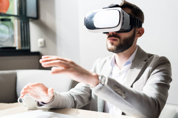 Бородатый бизнесмен в наушниках от виртуальной реальности сидит за столом и жестикулирует
 - Фото, изображение