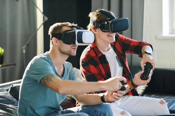 zwei junge Männer sitzen auf der Couch und spielen ein Spiel mit Virtual-Reality-Headset und Joypad - Foto, Bild