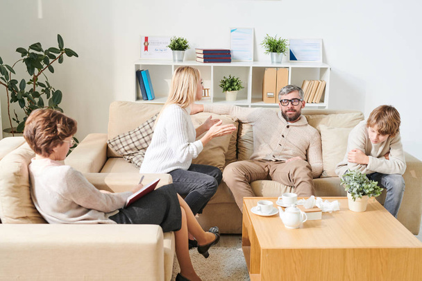 Stuffy young mother with blond hair sitting on sofa and gesturing hands while speaking claims to father at therapy session, sad son listening to quarrel of parents - 写真・画像