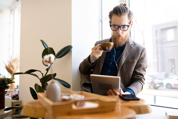 Serious concentrated hipster young man in glasses sitting on window-sill and listening to music in earphones while reading online book and drinking coffee in cafe. - 写真・画像