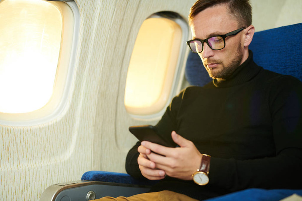 Портрет красивого взрослого мужчины с помощью смартфона, читающего электронные книги во время длительного перелета в самолете, копировальное пространство
 - Фото, изображение