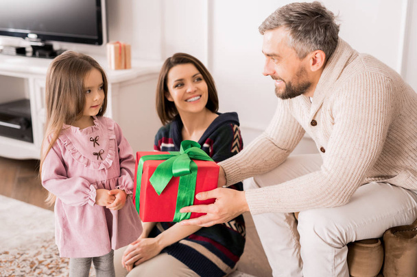 Портрет счастливого зрелого отца, дарящего подарок маленькой девочке, сцена, где семья обменивается подарками накануне Рождества
 - Фото, изображение
