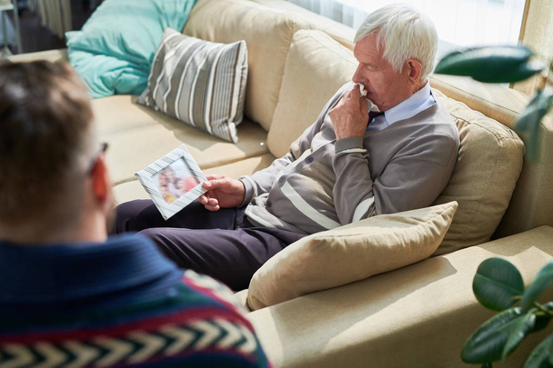 Despaired starszy mężczyzna w kardigan siedzi na kanapie i trzymając zdjęcie w ramce, dzieląc się jego problemy rodzinne z psychiatra na sesję terapii - Zdjęcie, obraz