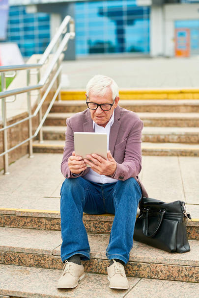 Ηλικιωμένος άνθρωπος σε λιλά μπουφάν χρησιμοποιώντας σύγχρονο tablet, ενώ κάθονται σε σκάλες δρόμο κοντά τσάντα - Φωτογραφία, εικόνα