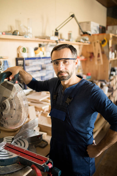 Waist up portrait of mature carpenter posing in workshop, standing next to electric disk saw - Fotó, kép