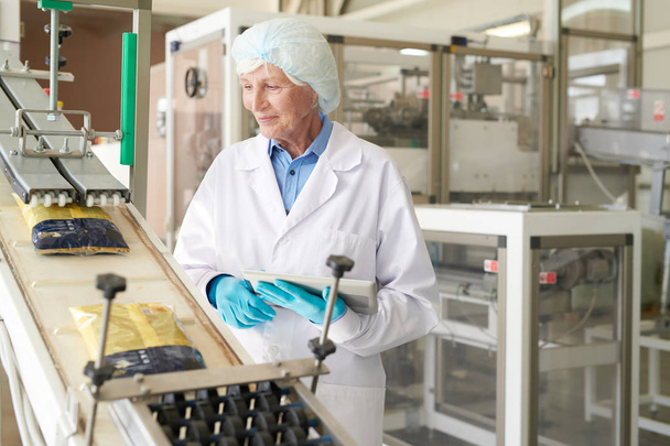 Portret uśmiechniętej Starsza kobieta kontroli procesu produkcyjnego w nowoczesnej fabryce żywności i patrząc na torby makaronu przesuwne w dół przenośnika taśmowego, przestrzeń kopiowania - Zdjęcie, obraz