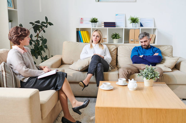 ernsthafte junge Paare in lässiger Kleidung sitzen auf einem bequemen Sofa und sprechen mit Psychologen, während sie ihre Beziehungsprobleme während der Sitzung lösen - Foto, Bild