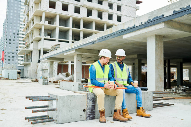 Serious content young multi-ethnic construction coworkers sitting on concrete beam with steel bars and chatting while drinking coffee during break - Photo, image