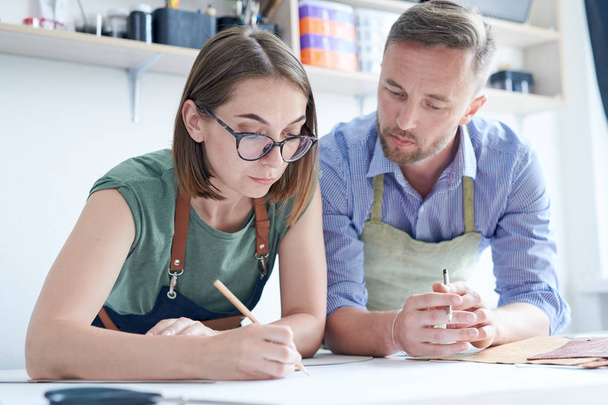 Jonge geconcentreerde vrouw tekent nieuwe schets van het product en toont deze aan haar partner aan tafel in de werkplaats - Foto, afbeelding