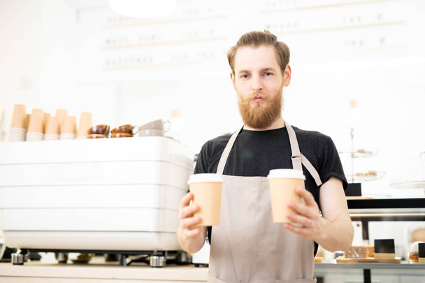 Kahve dükkanında duran ve müşteriye paket kahve fincanları verirken kameraya bakan apron içerik emin barista adam - Fotoğraf, Görsel