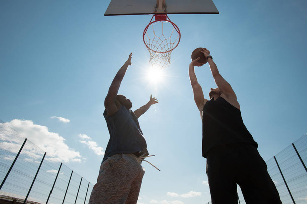 Visão de ângulo largo de dois homens jovens jogando basquete na quadra ao ar livre, foco no jogador atirando slam dunk, espaço de cópia
 - Foto, Imagem