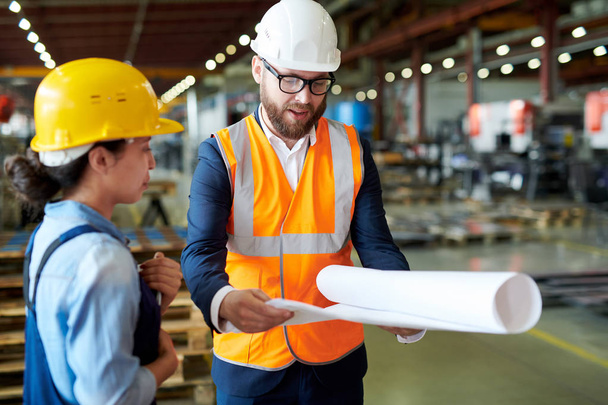 Porträt eines modernen bärtigen Ingenieurs mit Hut, der Baupläne hält, während er mit Arbeitern in der Fabrik über die Produktion diskutiert, Kopierraum - Foto, Bild