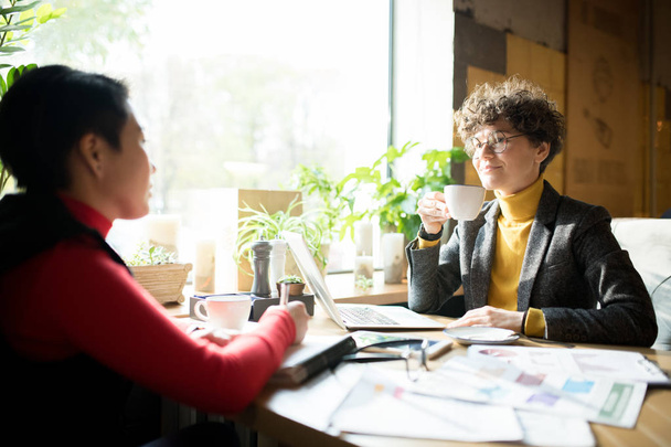Lächelnde selbstbewusste Business-Ladys in schicken Casual-Outfits sitzen mit Charts am Tisch und trinken Kaffee, während sie im Café über Marketing-Strategien diskutieren - Foto, Bild