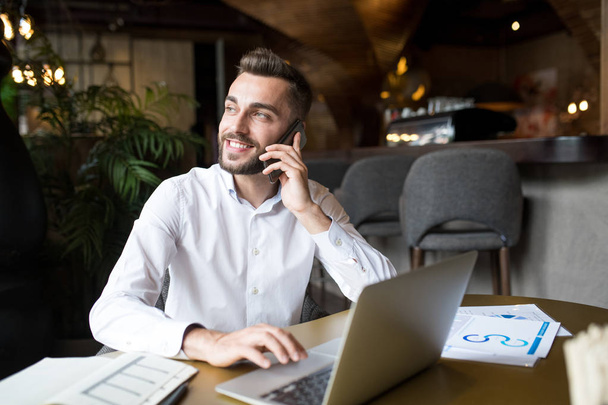 Portret młodego Brodatego biznesmen mówiący przez telefon i uśmiechnięty szczęśliwie podczas pracy z laptopem w kawiarni - Zdjęcie, obraz