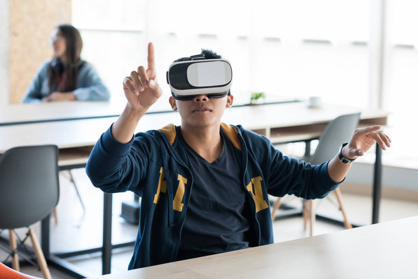Conteúdo curioso cara asiático moderno sentado à mesa e usando tela invisível enquanto testava o novo aplicativo de realidade virtual no escritório de espaço aberto
 - Foto, Imagem