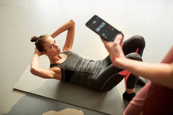 Над просмотром портрет современной молодой женщины, делающей хрусты во время фитнес-тренировки в оздоровительном клубе, в то время как женщина-инструктор держит смартфон с таймером
 - Фото, изображение