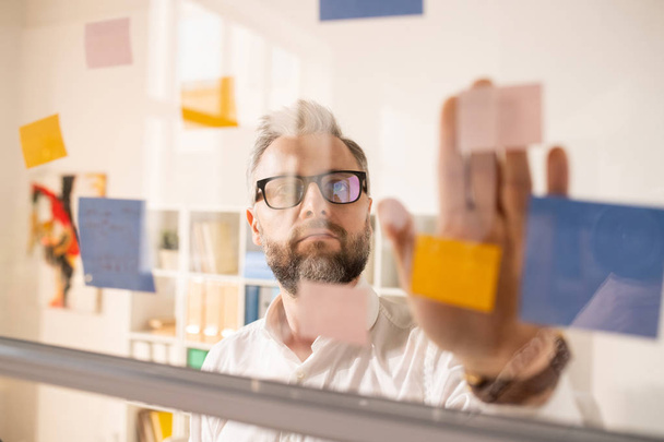 Empresário bonito introspectivo sério em óculos juntando adesivos em placa vítrea enquanto planeja novo projeto no escritório
 - Foto, Imagem