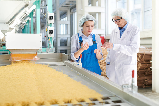 Portret dwóch kobiet omawiający produkcję stojącą przez przenośnik taśmowy w nowoczesnej fabryce żywności, przestrzeń kopiowania - Zdjęcie, obraz