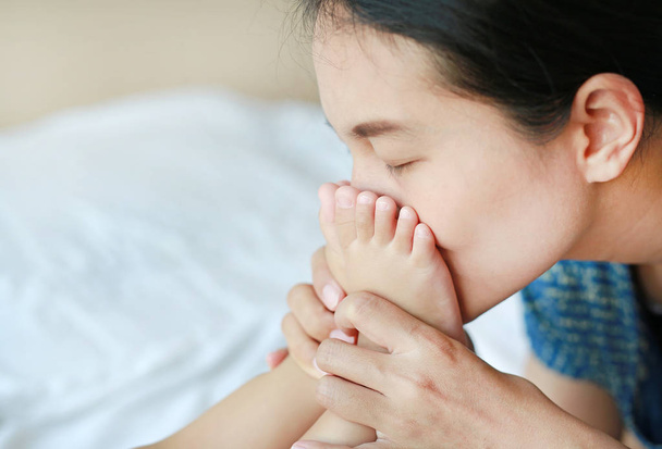 Peaceful Asian mother kissing baby feet on a white bed. - Foto, imagen