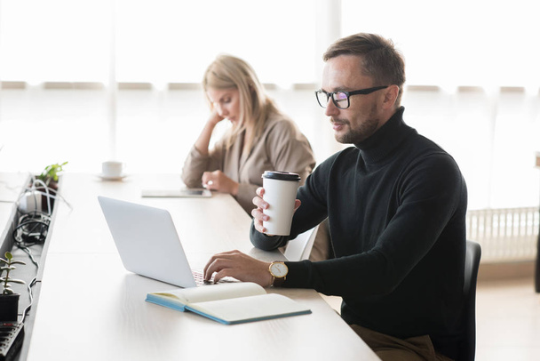 Grave pensativo jovem barbudo freelancer no pescoço pólo sentado à mesa e beber café ao usar laptop no espaço de co-trabalho
 - Foto, Imagem