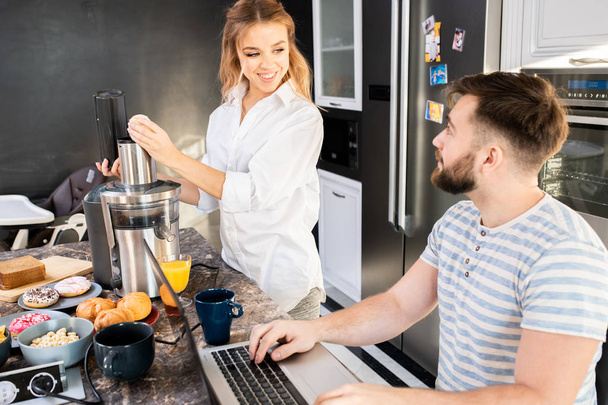 Portret van gelukkig jong stel thuis in de ochtend, focus op mooie vrouw koken in de keuken met man werken bij laptop naast haar, kopieer ruimte - Foto, afbeelding
