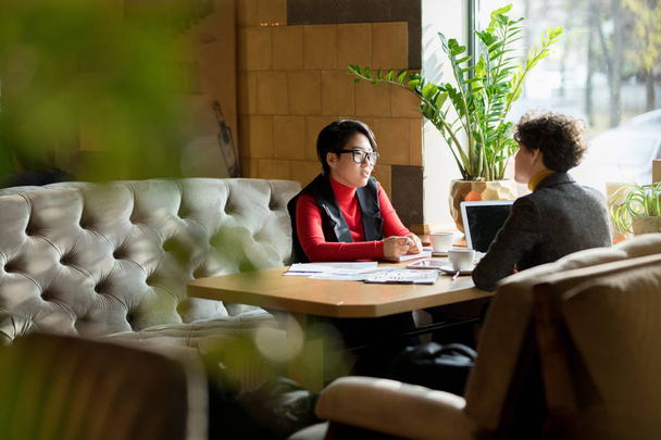 ernsthafte geschäftige multiethnische Damen in schicken lässigen Outfits sitzen einander am Tisch gegenüber und unterhalten sich über die Geschäftsentwicklung, während sie im modernen Café gemeinsam Berichte analysieren. - Foto, Bild