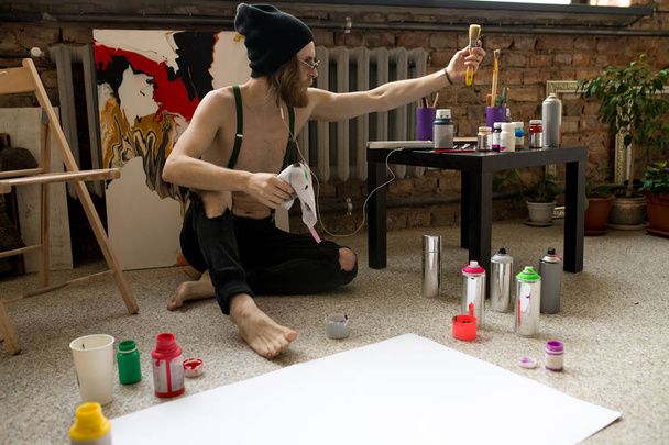 Täyspitkä muotokuva nykyajan mies taiteilija maalaus kuva istuu lattialla taidestudiossa, kopioi tilaa
 - Valokuva, kuva