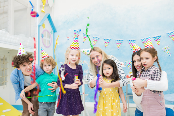 Позитивное возбуждение многонациональных детей с пустыми крекерами глядя на падение конфетти любопытно, их матери обнимают их на детской вечеринке
 - Фото, изображение