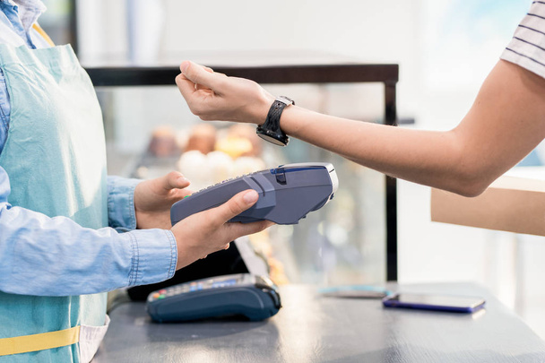 Side view mid section of unrecognizable woman paying by smart watch via NFC for her order in shop or cafe - Foto, afbeelding