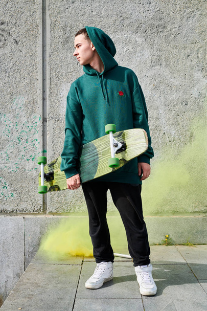 Full length portrait of modern teenager posing with longboard standing against concrete wall in green smoke - Photo, Image