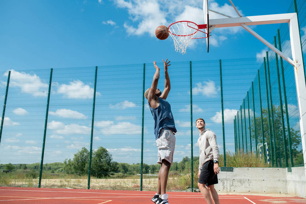 Vista de ângulo largo de dois homens jovens que jogam basquete na quadra ao ar livre, foco no homem africano atirando slam dunk em primeiro plano, espaço de cópia
 - Foto, Imagem