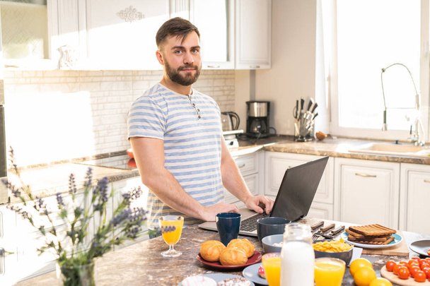 Portrait taille haute d'un bel homme barbu utilisant un ordinateur portable dans la cuisine tout en se tenant au comptoir pendant le petit déjeuner et en regardant la caméra, copier l'espace
 - Photo, image