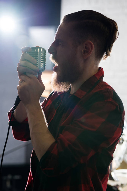 Πλευρική όψη με οπίσθιο φωτισμό πορτρέτο του σύγχρονου άντρα με γένια που τραγουδά στο μικρόφωνο ενώ εκτελεί στη σκηνή - Φωτογραφία, εικόνα