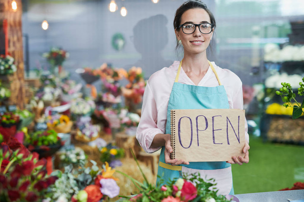 Portrait of modern young woman holding handwritten OPEN sign and smiling at camera while working in small flower shop, copy space - Photo, Image