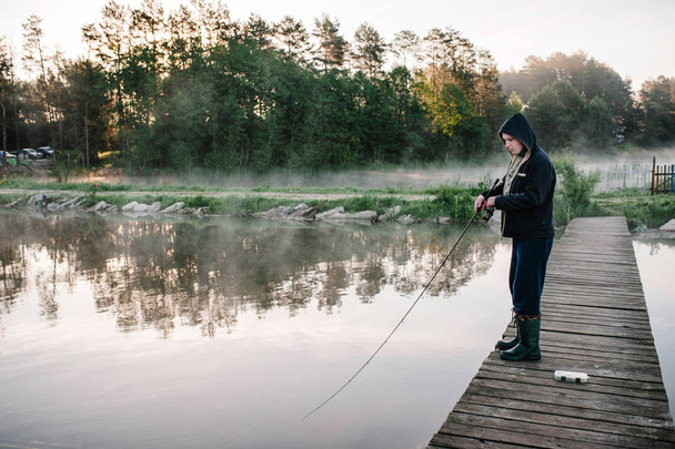 Fisherman with fishing rod standing on wooden bridge in misty morning  - Photo, Image