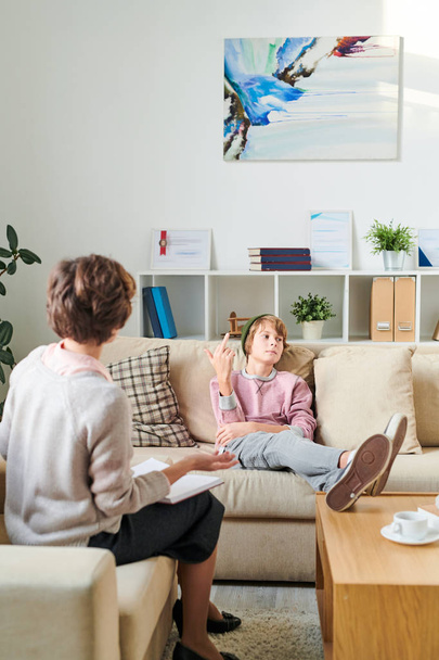 Serious indifferent teenager in hat sitting on sofa and keeping feet on table while fucking off psychiatrist at therapy session - Foto, Bild
