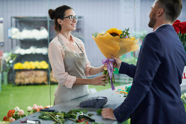 Side view portrait of smiling female shopkeeper handing bouquet to handsome gentleman buying flowers for date or celebration, copy space - Photo, Image