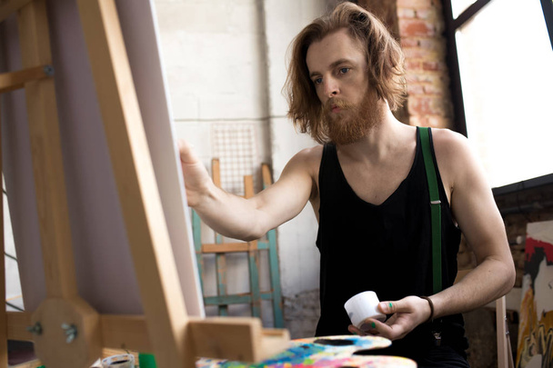 Πορτρέτο του εμπνευσμένη μακριά μαλλιά καλλιτέχνης κρατώντας βούρτσα και ζωγραφική σε καβαλέτο σε στούντιο τέχνης, αντίγραφο χώρου - Φωτογραφία, εικόνα