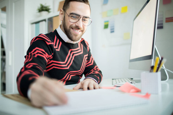 Веселий ініціативний красивий бородатий менеджер в окулярах сидить за столом з комп'ютером і записує ідеї під час роботи в сучасному офісі
 - Фото, зображення