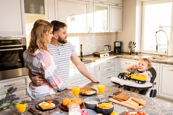 Muotokuva modernin perheen nauttia aamiaista keittiössä, nuoret vanhemmat etsivät söpö vauva istuu lapsi tuoli, kopioi tilaa
 - Valokuva, kuva
